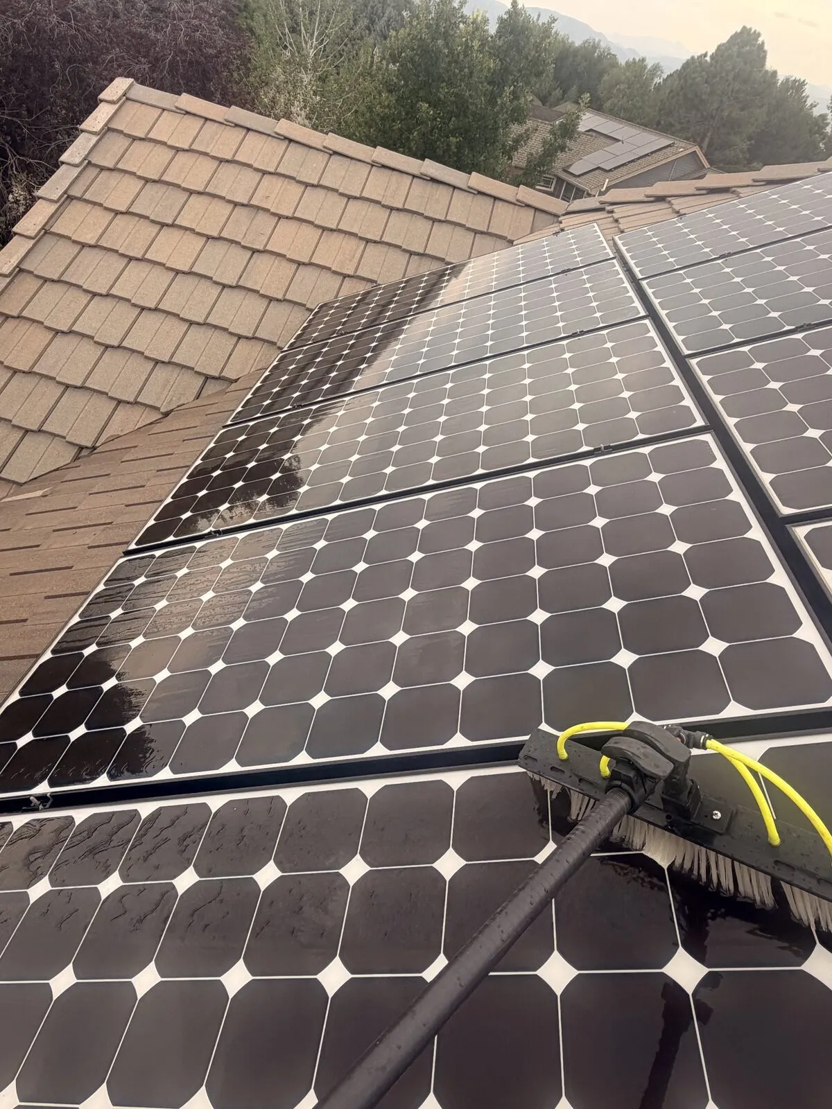 Water-fed pole brush cleaning rooftop solar panels on a tile-roof home in Boulder, Colorado, with cleaned panels visibly clearer than the uncleaned ones above